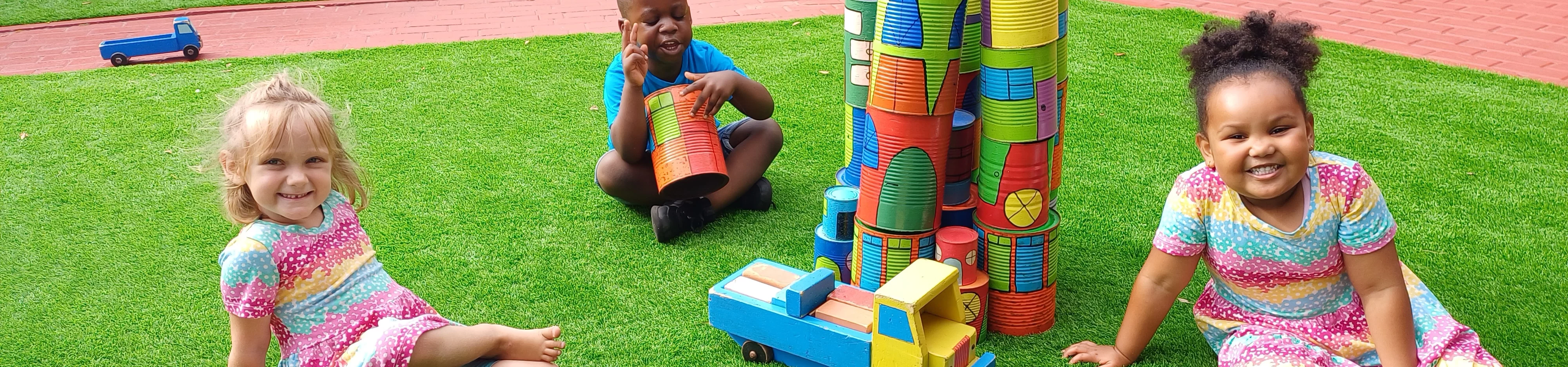 Three children sitting on the grass, engaged in play with colorful toy blocks scattered around them.
